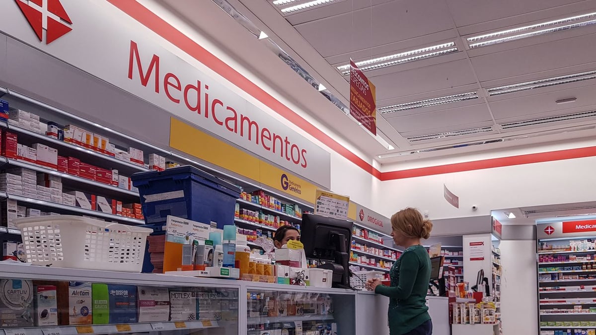 Cliente é atendida no balcão de uma farmácia, com prateleiras de medicamentos ao fundo (imagem ilustrativa)